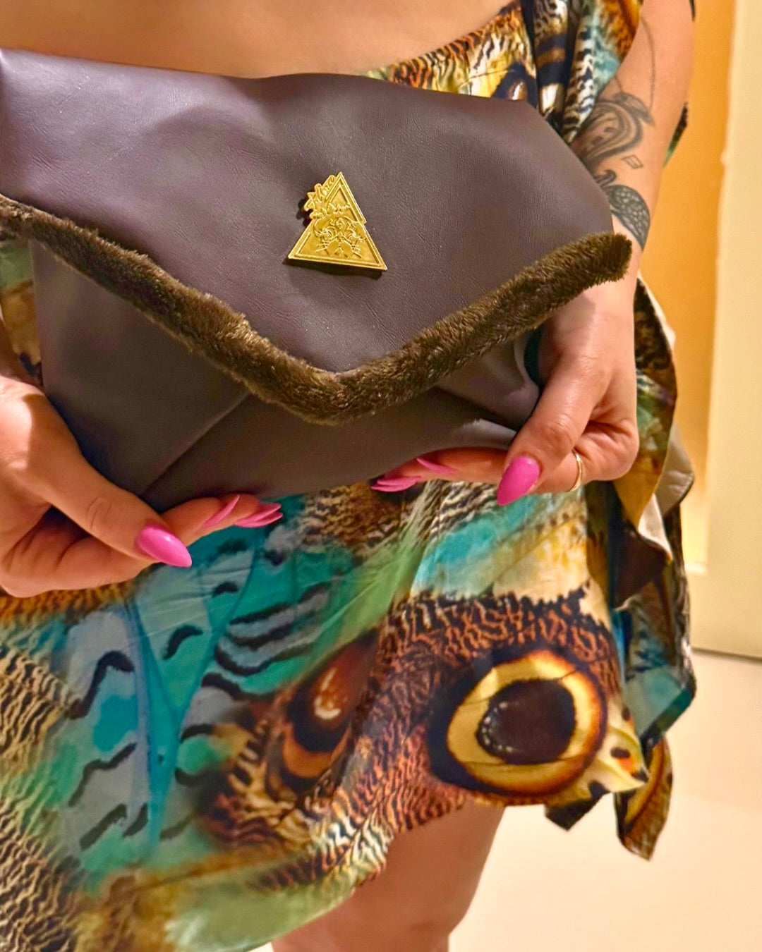 A close-up of a person holding a dark brown envelope-style clutch bag with a fuzzy trim and a small gold triangular logo. The person is wearing a colorful owl-patterned dress and has pink fingernails.