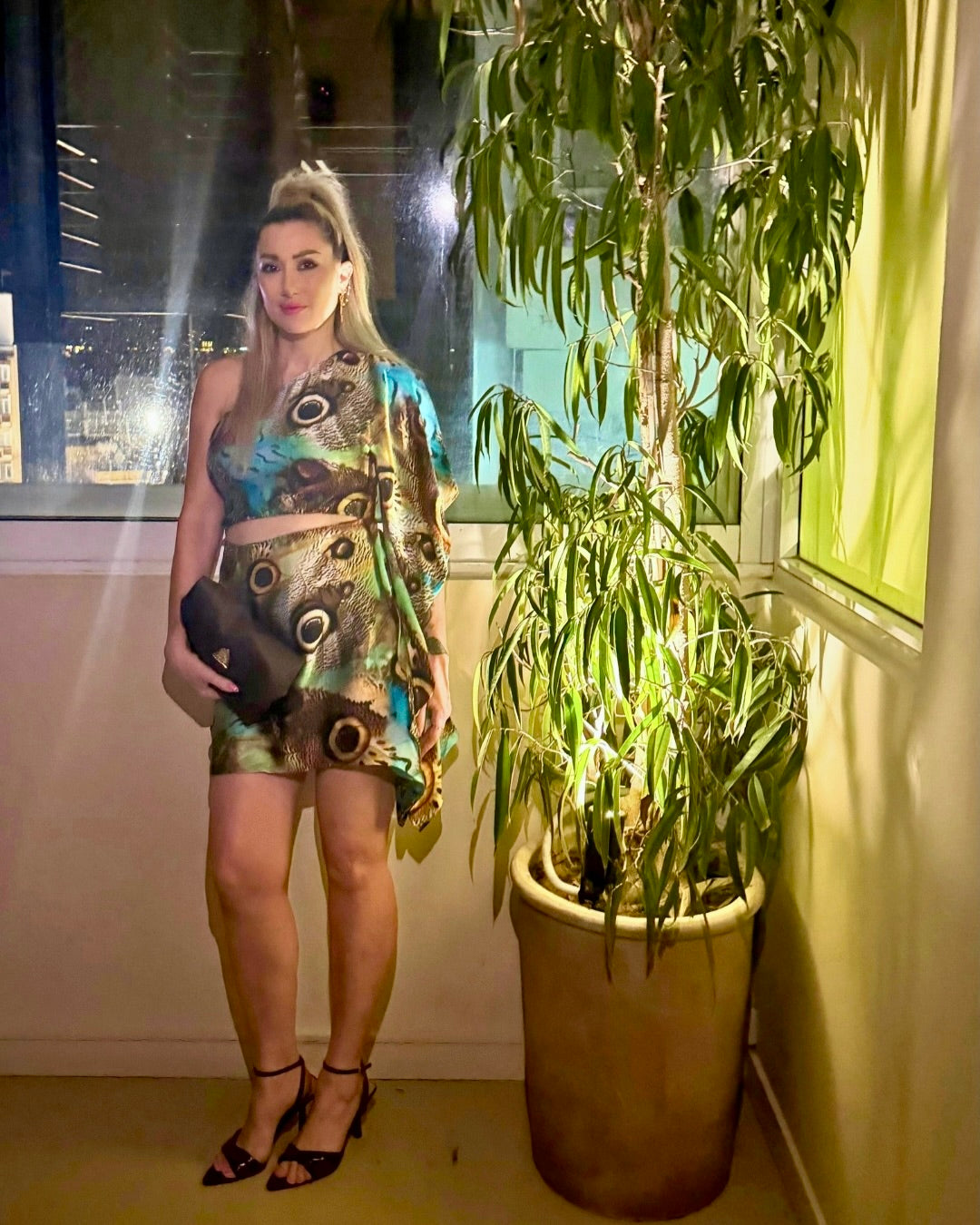 A woman stands indoors next to a large potted plant, wearing a matching two-piece outfit with a colorful abstract print in blue, green, and brown tones. She is holding a black clutch and wearing black strappy heels.