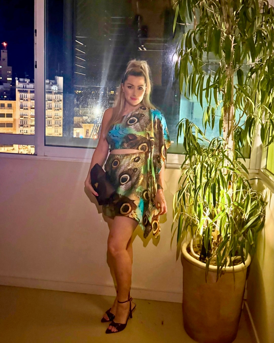A woman posing on a balcony at night wearing a colorful, patterned two-piece set consisting of a halter crop top and a matching mini skirt. She is holding a black clutch and wearing black high-heeled sandals.
