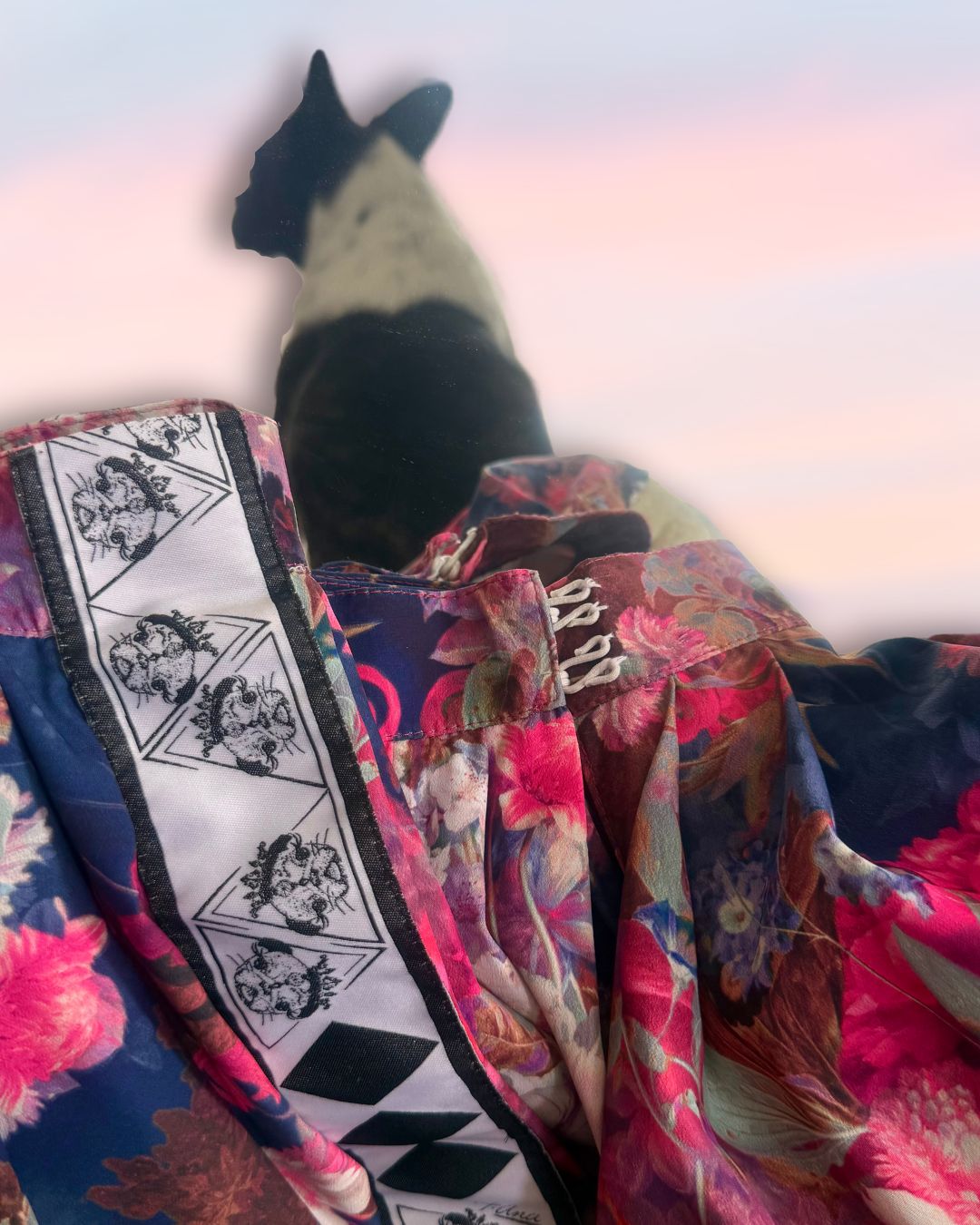 Luxury detail of High-quality floral fabric detail showing print and texture of couture three-piece set; a floral satin couture set in blush and fuchsia tones by t•Ana Couture; captured under Mediterranean sunlight with divine flow and effortless elegance. Designer logo close-up details and dog muse in the background. 