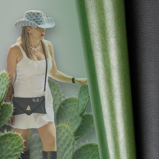 Woman wearing black Desserto cactus leather clutch bag by t•Ana Couture styled in nature with cactus plants and sustainable plant-based leather material. Woman in a white dress and hat standing among cacti with a blurred green and black fabric in the foreground. 