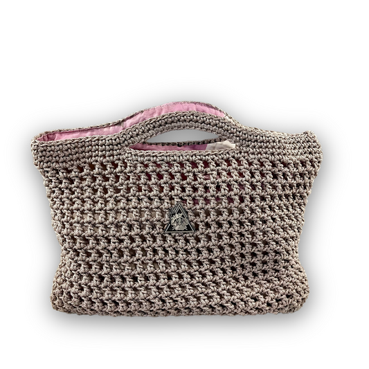 Crocheted handbag with a brand logo held over a branded surface. Mystic Mocha Bloom hand knit mocha handbag by t.Ana Couture; artisan crafted beige grey knit bag with organic linen zinnia floral lining from Mediterranean Muse Sand and Sea Collection
