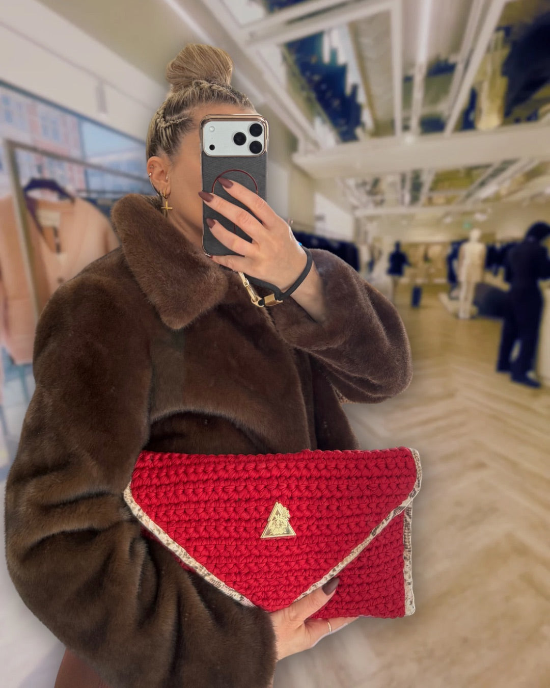 Luxury setting of glamorous elevated high value lady taking a selfie in a high end shop in london holding the Sacred Venom Magenta Knit Clutch Handbag – magenta red knit clutch with gold logo and beige snake-print lining from t•Ana Couture’s Mediterranean Muse: Sand & Sea Collection; symbolizing passion, transformation, and divine feminine grace.