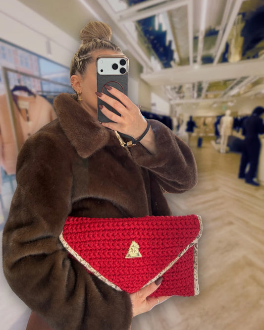 Luxury setting of glamorous elevated high value lady taking a selfie in a high end shop in london holding the Sacred Venom Magenta Knit Clutch Handbag – magenta red knit clutch with gold logo and beige snake-print lining from t•Ana Couture’s Mediterranean Muse: Sand & Sea Collection; symbolizing passion, transformation, and divine feminine grace.
